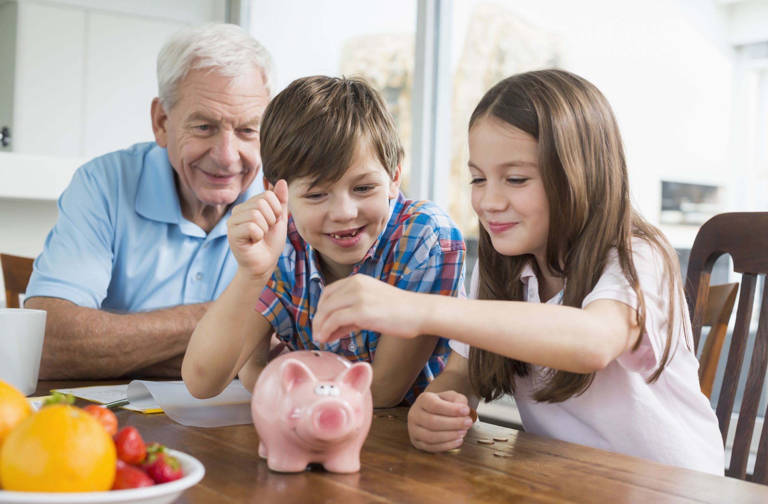 A grandfather watches two children put coins into a pink piggy bank on a table.