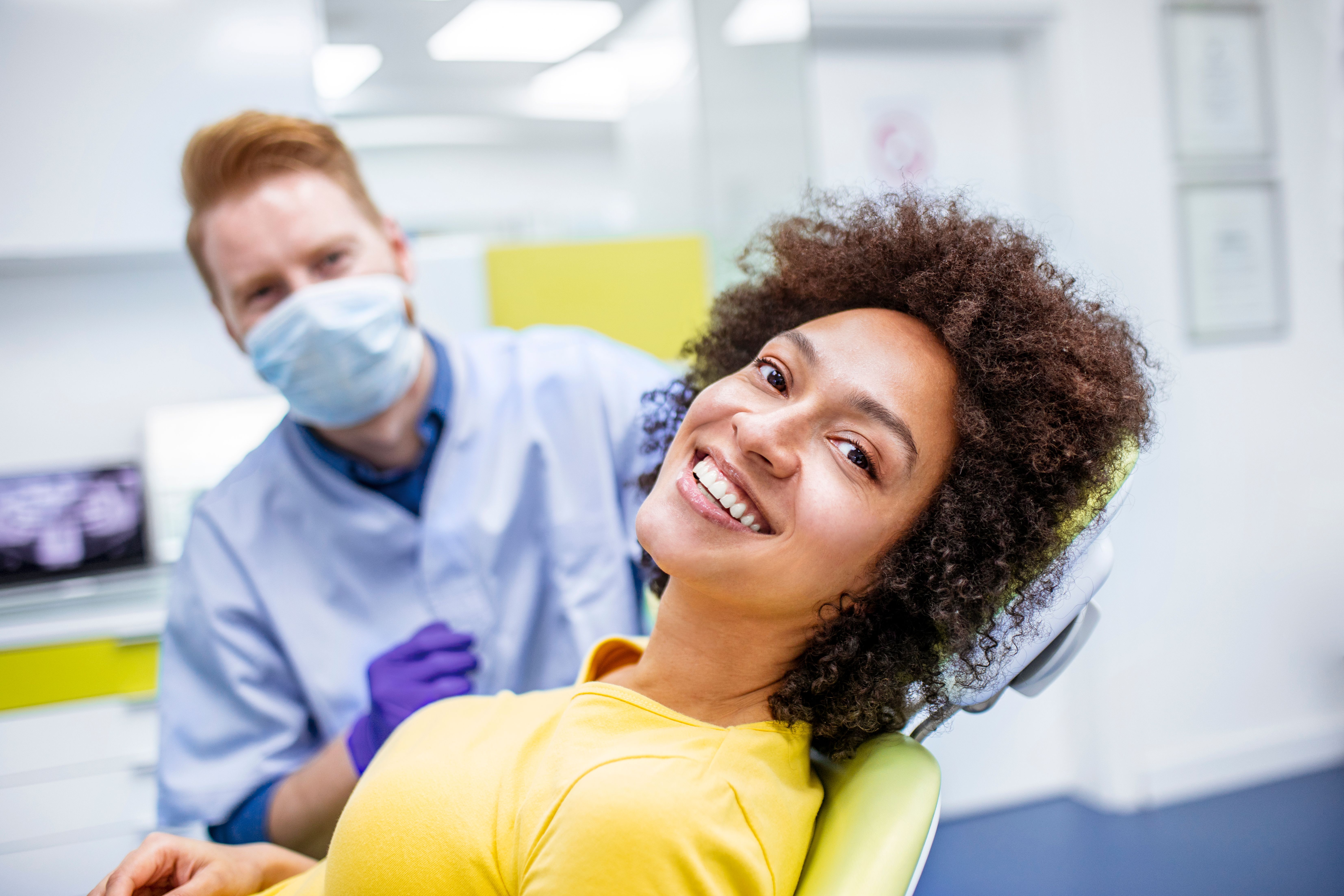 Smiling woman in a dental chair with a masked dentist behind her.