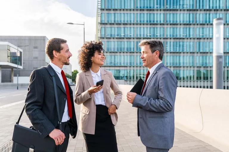 Three business professionals talking and smiling outdoors with modern buildings in the background.