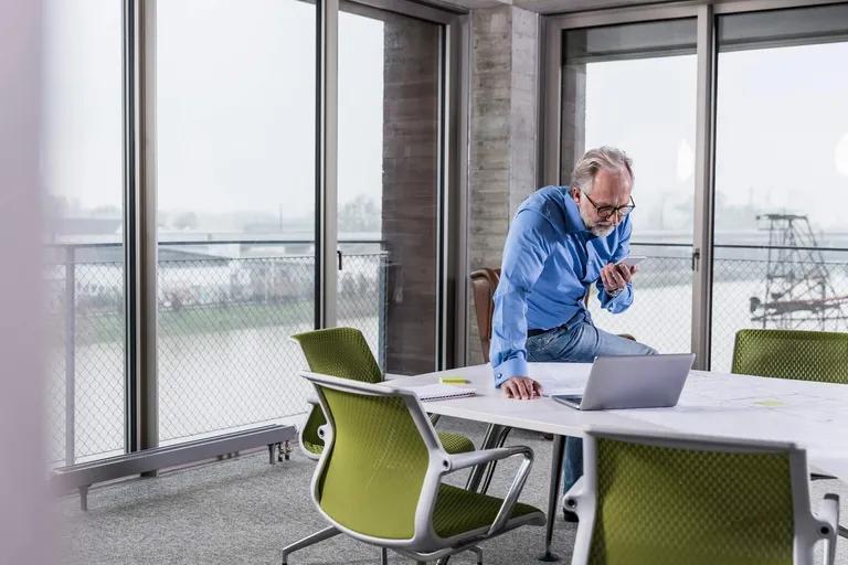 A senior man leaning on a conference table, looking at his smartphone, with a laptop nearby.