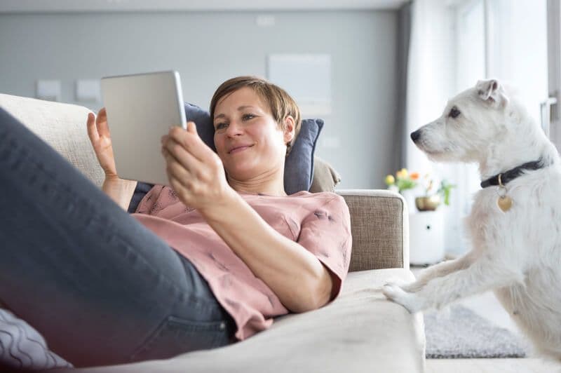 Eine Frau liegt auf einer Couch und benutzt ein Tablet, während ein Hund zuschaut.