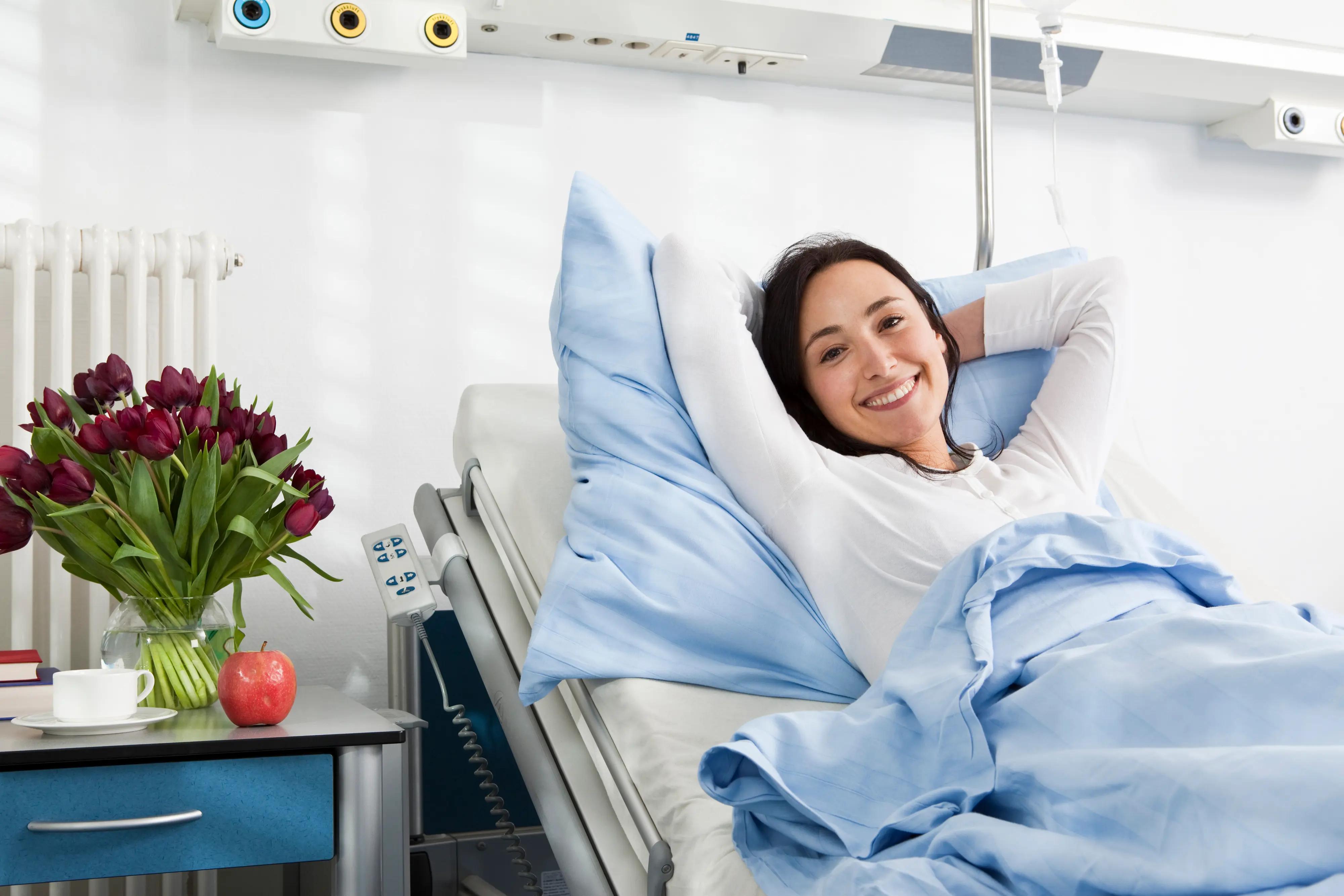 A smiling woman lies comfortably in a hospital bed with an IV pole nearby and flowers on a bedside table.