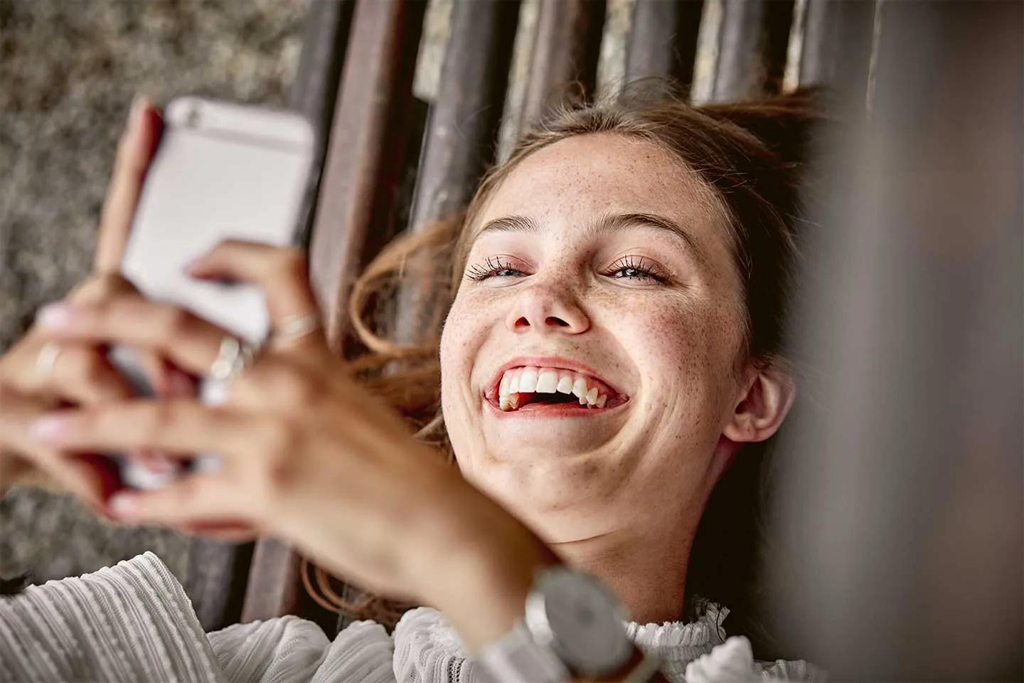 A joyful, freckled woman lies on her back, holding a smartphone.