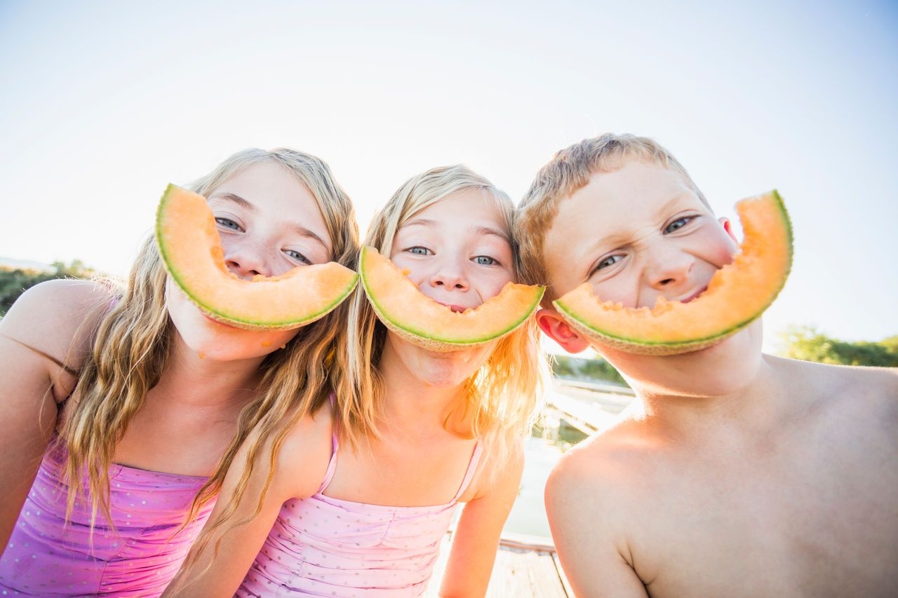 three children are holding slices of cantaloupe in front of their faces .