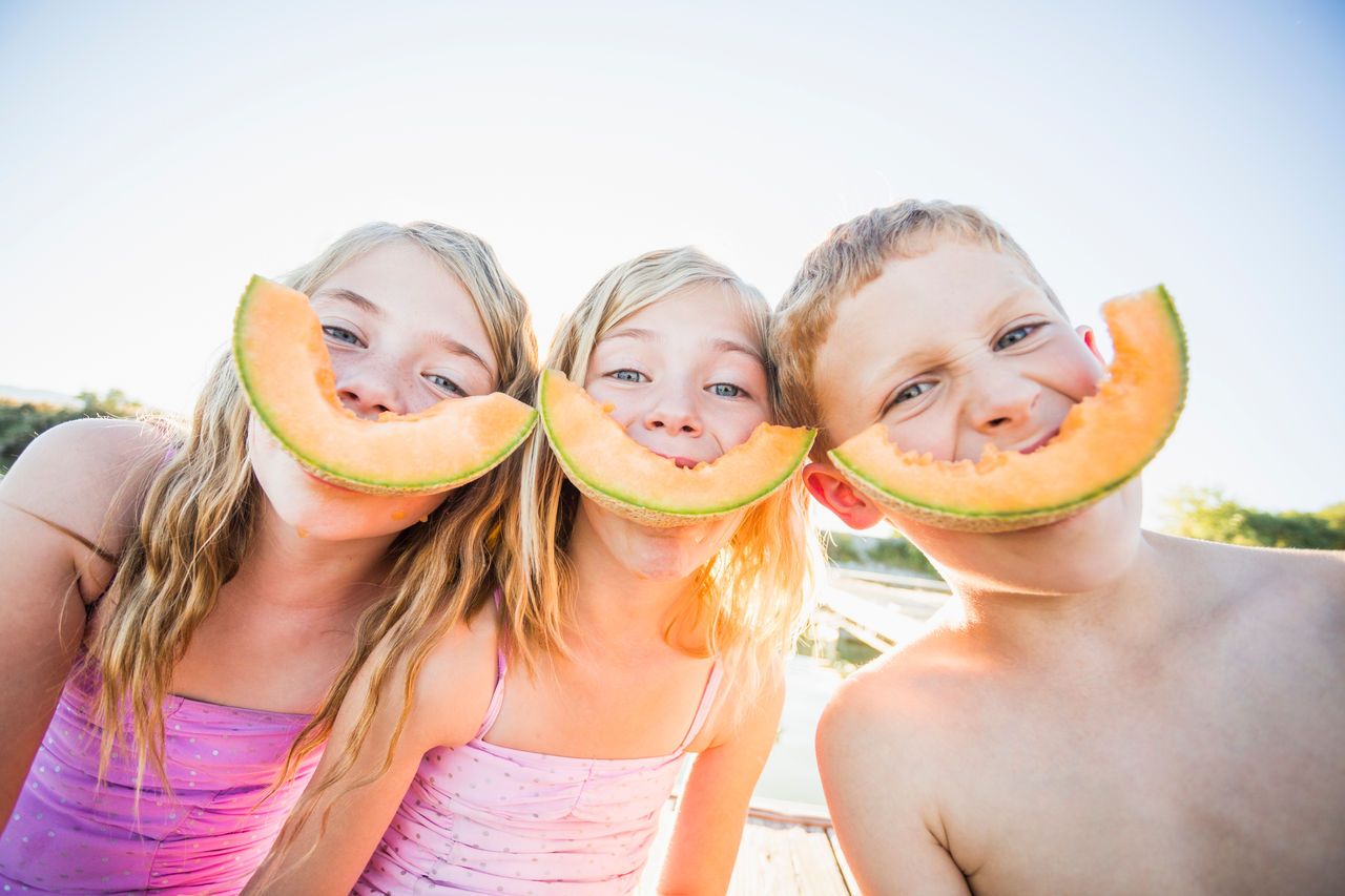 Three children playfully hold melon slices to their mouths like big smiles.
