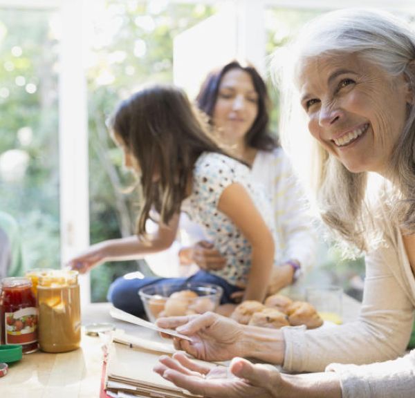 Smiling senior woman holding a recipe card, with a mother and child preparing food in a bright kitchen.
