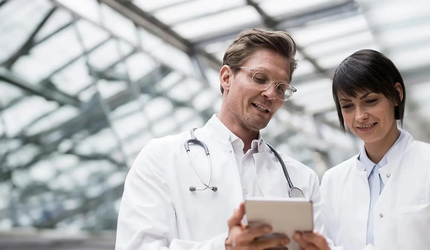 Two smiling doctors in lab coats looking at a tablet.