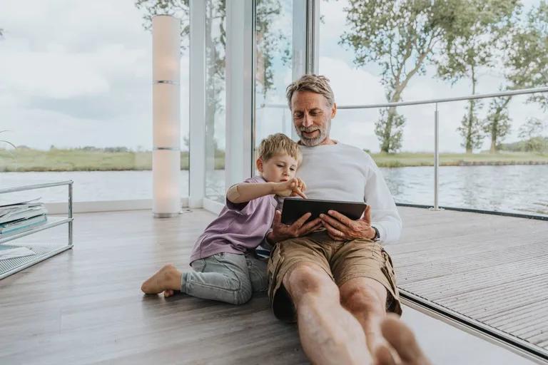 A man and boy sit on the floor by a large window overlooking water, interacting with a tablet.