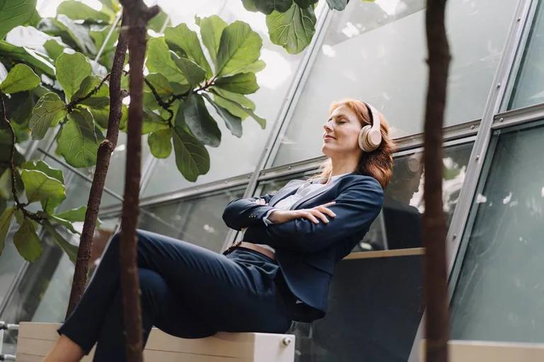 A woman in a suit wearing headphones, eyes closed, relaxing surrounded by plants.