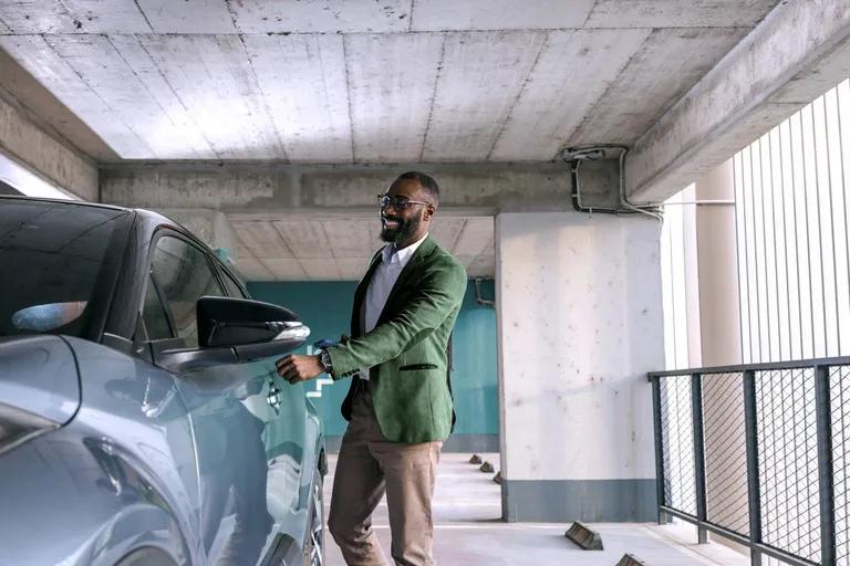 A smiling man in a green jacket opens a car door in a parking garage.