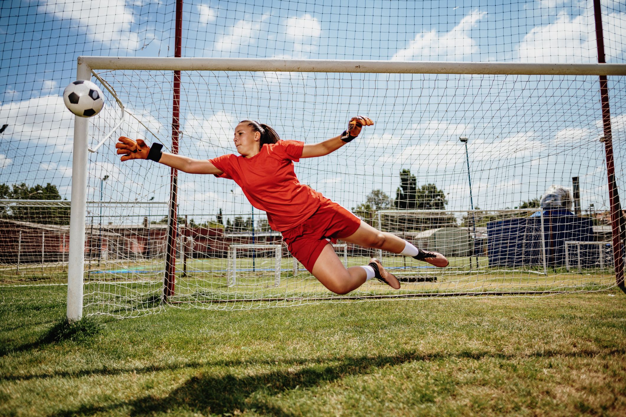 Female soccer goalkeeper in red uniform dives mid-air to block a soccer ball at the goal.