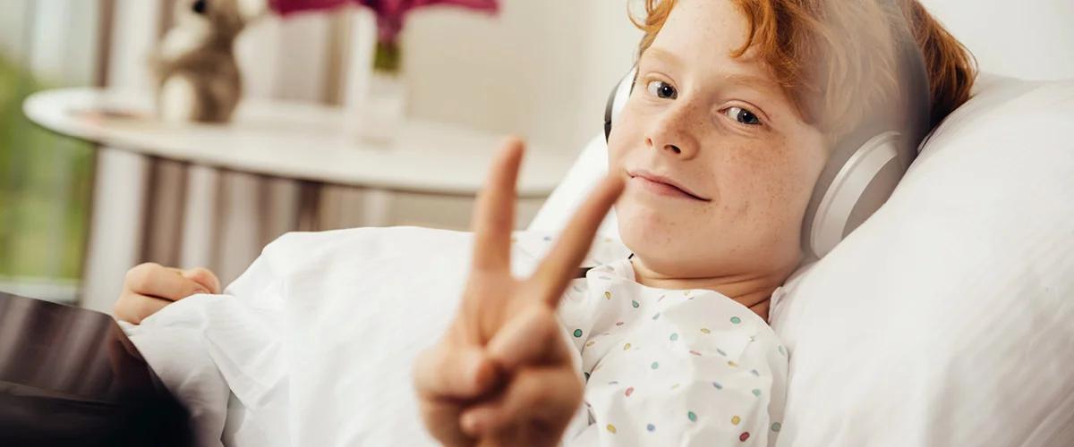 Red-haired child in bed wearing headphones, smiling and making a peace sign.