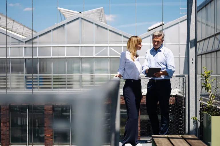 Two professionals on a rooftop reviewing a tablet with a modern glass building behind them.